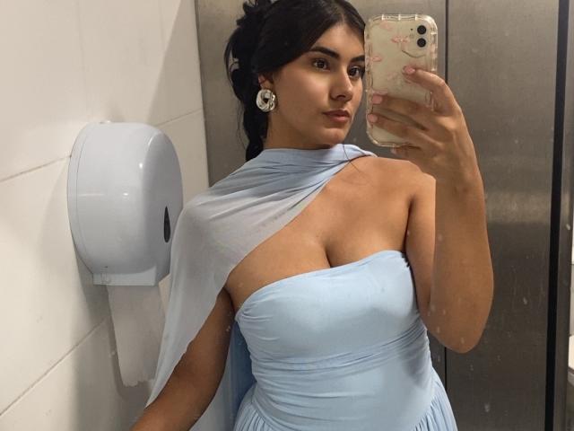 AlaraKey with big tits in a light blue dress, one-shoulder, showing cleavage, holding a phone, wearing white earrings, in an elevator, looking at the mirror.