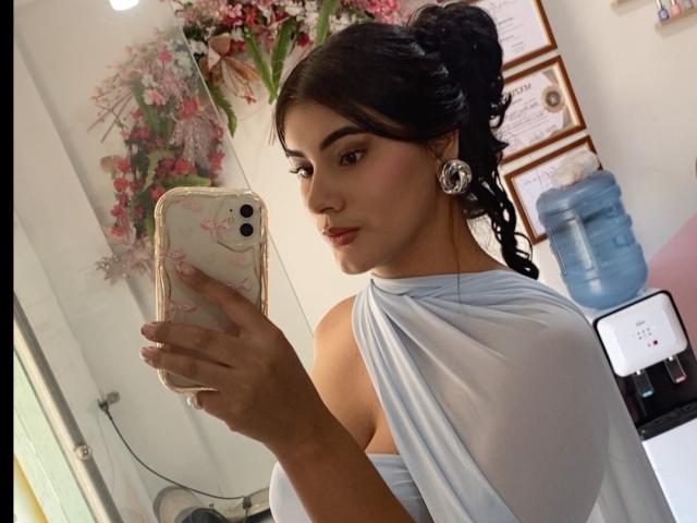 AlaraKey with dark hair in a bun, wearing a white off-shoulder top, taking a mirror selfie with a phone, showing her face and earrings, holding her phone in her hand.
