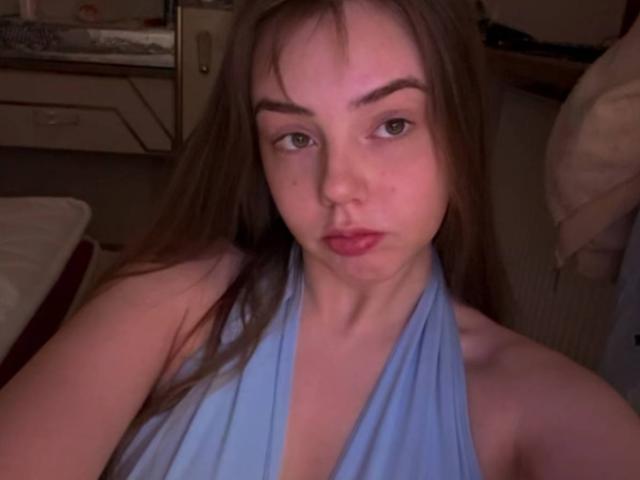 LauranTanton with long brown hair, blue halter top, pouty lips, and eyebrows slightly raised, looking at the camera with a neutral expression. Boobs are partially visible.