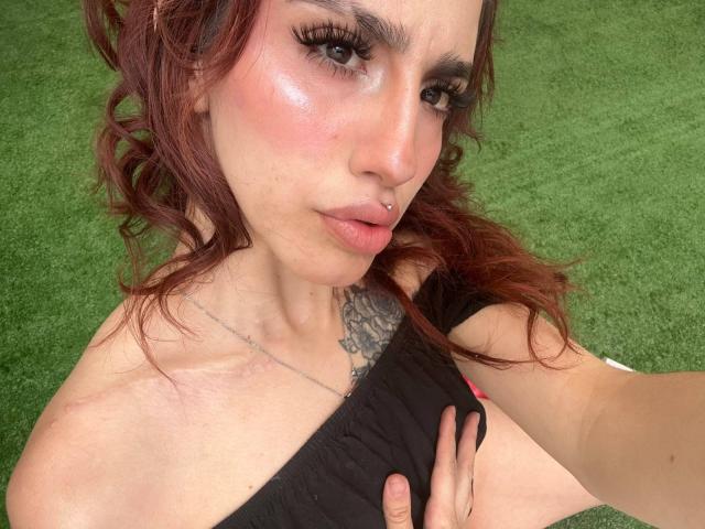 ElodieRoussee has curly red hair, a nose piercing, and a tattoo on her chest. She's holding her black top, showing off her tits and a hand on her chest.