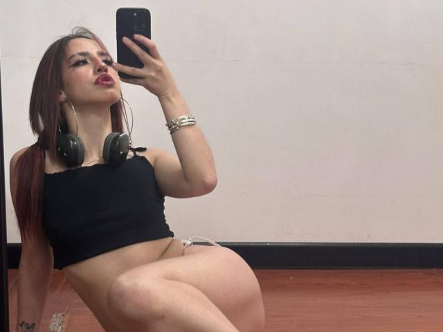 Elodie Roussee, avec ses cheveux roux, son haut noir sans bretelles, ses écouteurs et sa montre, prend un selfie devant un miroir, montrant ses seins et ses fesses, assise sur un plancher en bois..