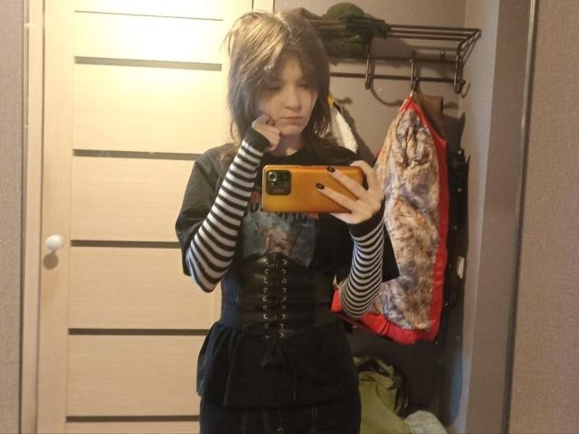 LinaMuller with shoulder-length brown hair, striped sleeves, black corset, and black shirt, holding orange phone, standing in mirror, looking serious, in a cluttered room.