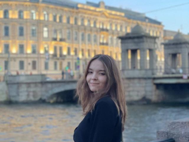 LinaMuller tem seios médios, cabelo castanho ondulado e um sorriso adorável, vestindo uma blusa preta, e está parada perto de um rio com um edifício de pedra histórico ao fundo, ao entardecer..