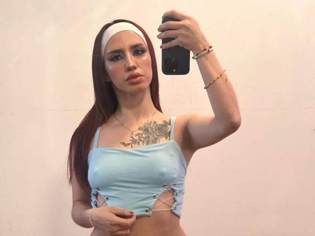 ElodieRoussee with red hair, white headband, light blue crop top showing small tits and a chest tattoo, taking a mirror selfie with her right arm raised.