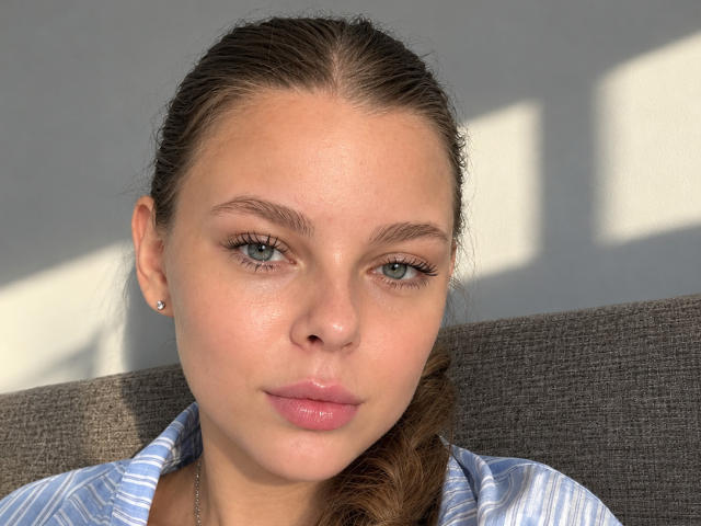 RhonaBells has fair skin, blue eyes, and pink lips. She's wearing a striped shirt, with a braid over her shoulder, and has a small stud earring in her left ear.