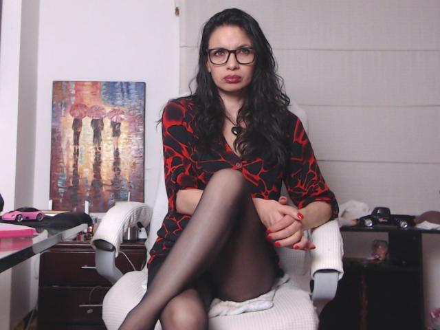 AfroditaRgenis, com cabelos pretos longos, óculos, vestido com padrão vermelho e preto, pernas cruzadas com meia-calça preta, esmalte vermelho, sentada em uma cadeira branca, olhando para a câmera..