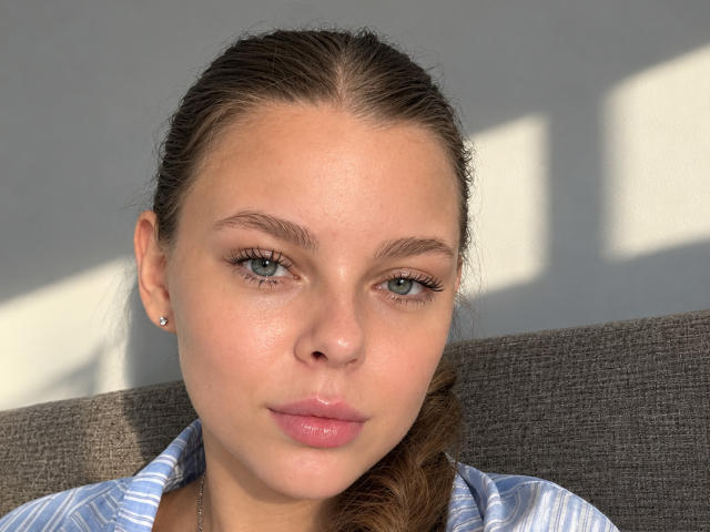 A RhonaBells tem um rosto adorável, com olhos azuis, lábios rosados e um leve franzido. Ela está vestindo uma camisa listrada, tem uma pequena argola no ouvido e seu cabelo castanho está preso para trás..