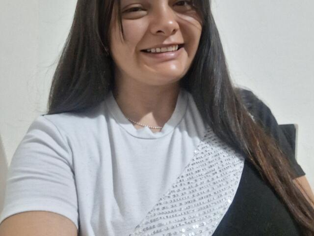 Katherin Latin tiene el cabello largo y oscuro, está sonriendo mostrando sus dientes, y lleva una camisa blanca y negra con lentejuelas plateadas. Lleva un collar pequeño..