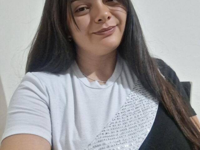 Katherin Latin ha i capelli lunghi e scuri, la pelle chiara e un leggero sorriso. Indossa una maglietta bianca con un motivo argentato e una borsa a tracolla nera..