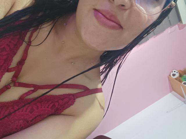 LunaWhitmore with wet black hair, glasses, red lace bra showing small tits, pink lips, and chest piercings, smiling in a pink room with a stuffed bear.