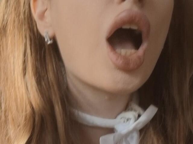 ElzaKomx's open mouth with slightly parted lips, showing teeth, and wearing a white choker with a bow. Her blonde hair and small earrings are visible.