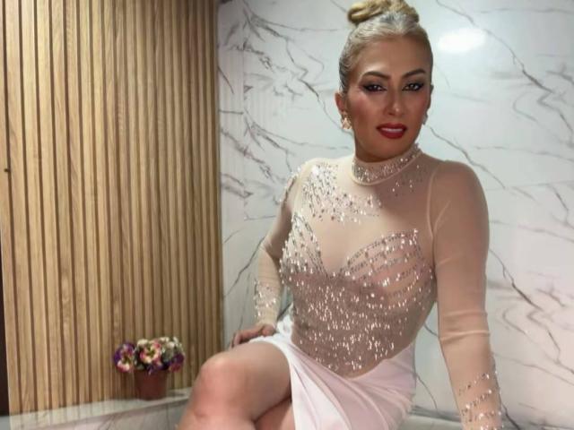 MisseScarleth com coque loiro, batom vermelho e blusa transparente e brilhante, mostrando os seios, sentada com saia branca, vaso de flores ao fundo, parede de madeira, parede de mármore..