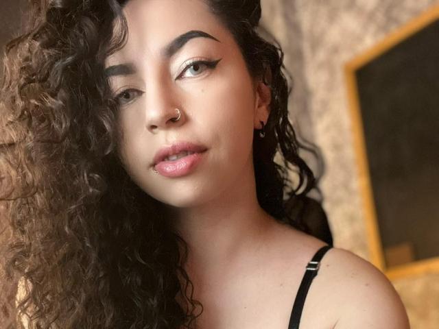 ScarlattaaDesire tiene el pelo castaño rizado, un piercing en la nariz y lápiz labial rosa. Lleva un top negro con tirantes y mira a la cámara con delineador de ojos alado. Su trasero no es visible..