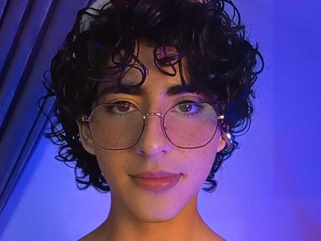 ElliotNoir, avec ses cheveux noirs bouclés, porte des lunettes rondes et un boucle d'oreille argentée. Il/Elle a des taches de rousseur et un léger sourire, sous un éclairage bleu. Ses seins ne sont pas visibles..