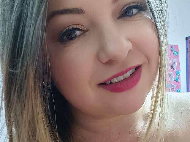 NatalyHami, com cabelo loiro, olhos azuis, batom rosa e dentes brancos, sorrindo para a câmera, mostrando seu rosto suave e arredondado e sua pele clara..