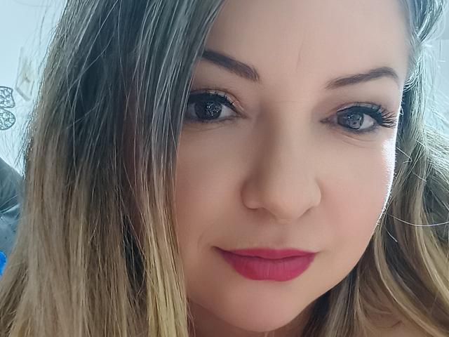 NatalyHami tiene el cabello rubio, ojos azules y lápiz labial rojo. Sus pestañas son largas y está mirando a la cámara con una ligera sonrisa. Su nariz está centrada..