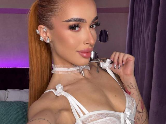 MarissaFex with orange ponytail, white lace lingerie, flower earrings, holding silver spoon to her pink pout, tattoos on arm, choker with pearl, purple curtains in background.