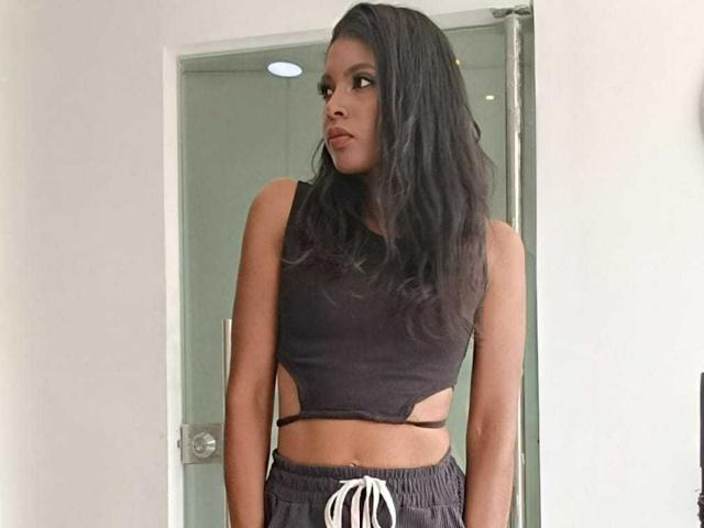 CamilaWest ha i capelli scuri e ondulati, la pelle marrone e indossa un top corto nero con aperture, che lascia intravedere la parte centrale del suo addome, e pantaloni neri con un cordoncino bianco..