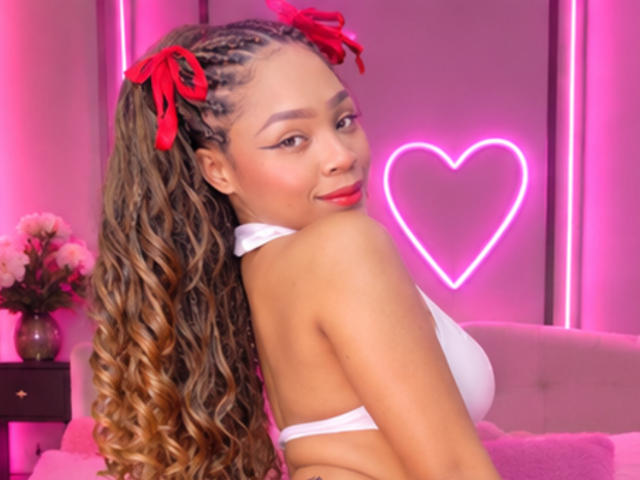 SkinnySamantha con lunghi capelli castani ricci, fiocchi rossi per capelli, top di bikini bianco, cuore neon rosa e rossetto rosso, sorride guardando oltre la spalla, mostrando il seno, in una stanza illuminata di rosa..