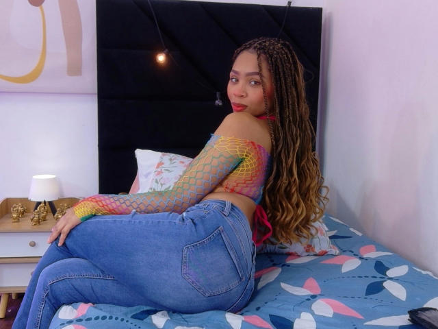 SkinnySamantha, con lunghi capelli intrecciati, una maglietta di rete colorata e jeans blu, seduta su un letto, che guarda oltre la spalla, mostrando il suo sedere. Il letto ha una coperta blu con motivi..