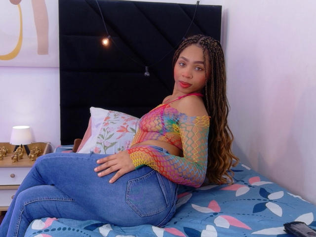 SkinnySamantha with long braids, colorful fishnet top, and blue jeans, sitting on a bed with a patterned blue blanket, showing her ass and tits, with a cute red lip.