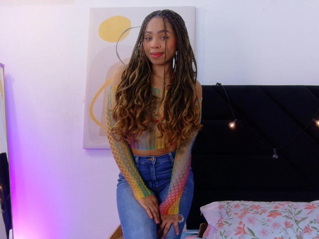 SkinnySamantha with long, wavy brown braids, colorful mesh top, and blue jeans, standing with a slight smile, hands near her crotch, in a brightly lit room.