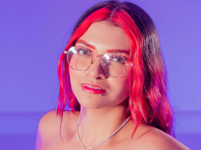 AriaRosel with pink and black hair, wearing clear glasses and a silver necklace, has shiny pink lipstick and a small nose ring, set against a purple background.
