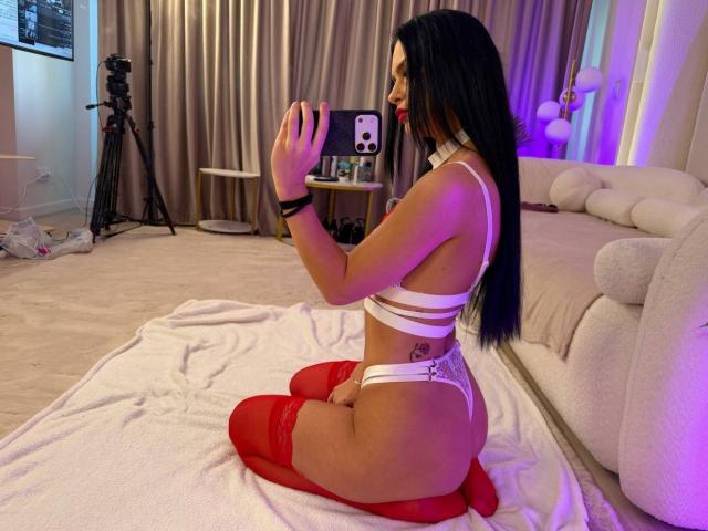 IliriaKara with long black hair, kneeling on a towel, wearing white strappy lingerie, red thigh-highs, and red lipstick, holding a camera, showing her ass and tattoo.