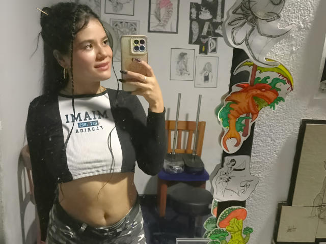 IsabellaVoss with long black hair, white and black raglan shirt, and shiny silver pants, holding a phone, mirror selfie, smiling, tattoos on wall, fish sticker.