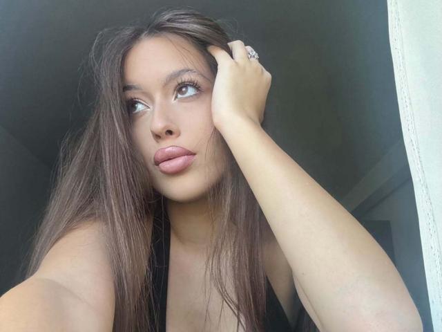 BrooklynChase with long brown hair, pouty pink lips, and a ring on her right hand, looking up with her left hand in her hair. She's wearing a black top.