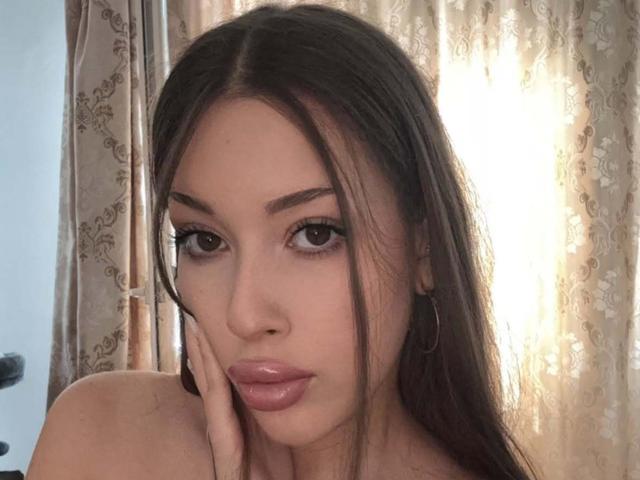 BrooklynChase has long brown hair, full pink lips, and brown eyes with thick eyelashes. She's touching her cheek with her hand, wearing a hoop earring.