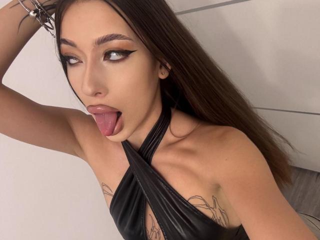 BrooklynChase has long brown hair, dark eyeliner, and is sticking out her tongue while wearing a black halter top, showing tattoos on her chest and a bracelet on her wrist.