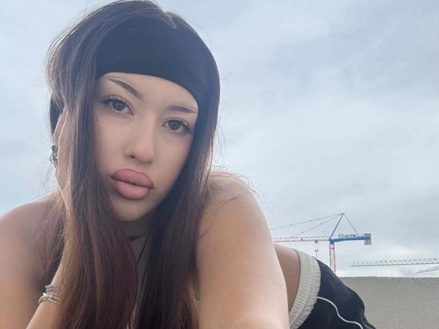 BrooklynChase with long brown hair, black headband, and pink lips, leaning forward, showing her tits, in a sleeveless top, with a crane in the blurry background.