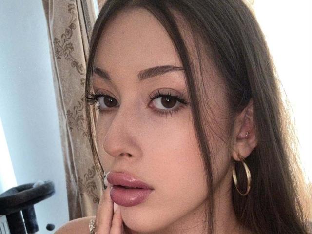 BrooklynChase has pouty pink lips, brown eyes with long lashes, and brown hair, wearing gold hoop earrings and a small nose piercing, with a finger on her chin.