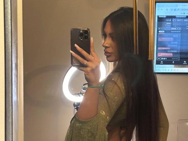 CristalSky, con cabello largo y castaño oscuro, una blusa verde bordada, sosteniendo un teléfono negro, tomando una selfie frente al espejo, mostrando su perfil y su trasero ligeramente visible. Una luz de aro en el fondo..