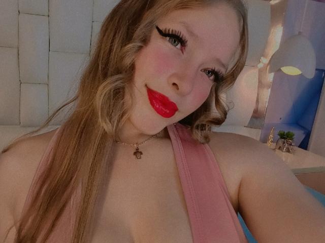 IvyBellmont, con lunghi capelli biondi, rossetto rosso e una maglia rosa che mette in mostra il décolleté, con l'eyeliner nero e una collana d'oro, sorride alla fotocamera..