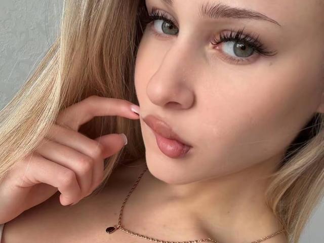 MilagrosEdmisten's face, blonde hair, green eyes, full lips, light skin, pink nails, touching her lips with her right hand, wearing a gold necklace with a small heart pendant.