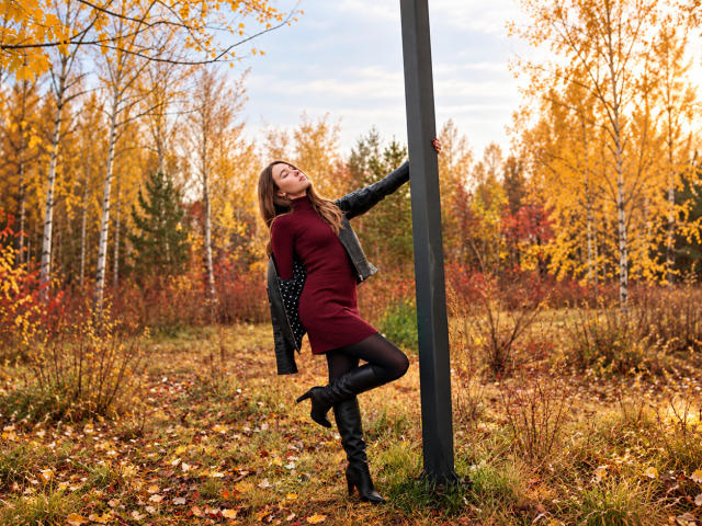AmmaLoyXX stands against a metal pole, wearing a red dress, black boots, and a gray jacket, with autumn leaves and trees in the background. Her ass is slightly raised.