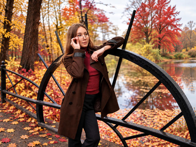 AmmaLoyXX, with long brown hair, brown coat, red sweater, and black pants, leans on a black wheel, talking on a cellphone, surrounded by autumn leaves and a pond.