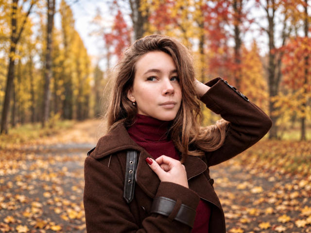 AmmaLoyXX con abrigo marrón, cuello alto rojo y mochila negra, con la mano en el pelo, bosque otoñal, hojas amarillas y rojas, mirando hacia un lado, busto mediano, linda, luz natural..