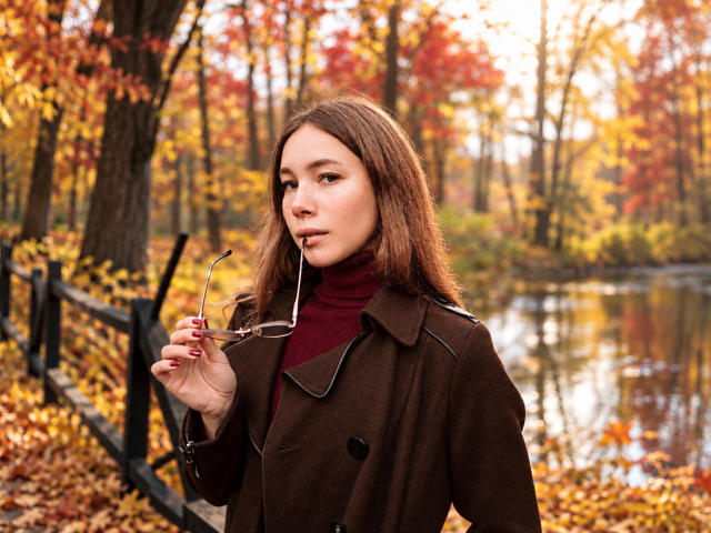 AmmaLoyXX with brown hair, red turtleneck, and brown coat, holding glasses in autumn forest, reflection in water, tree and fence in background, looking slightly to the side.