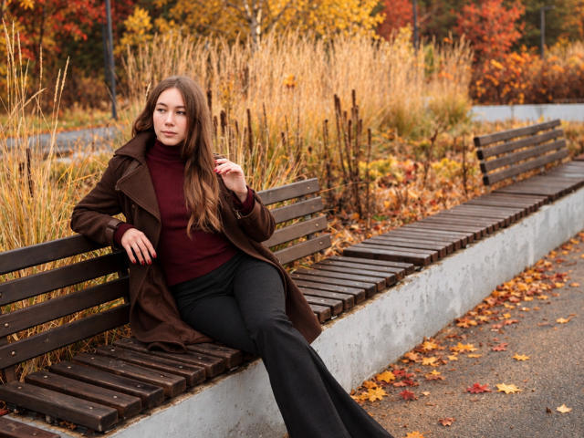 AmmaLoyXX, con el pelo largo y castaño, está sentada en un banco de madera en otoño, vestida con un abrigo marrón, un cuello alto rojo y pantalones negros, con la mano cerca de la cara, luciendo relajada..