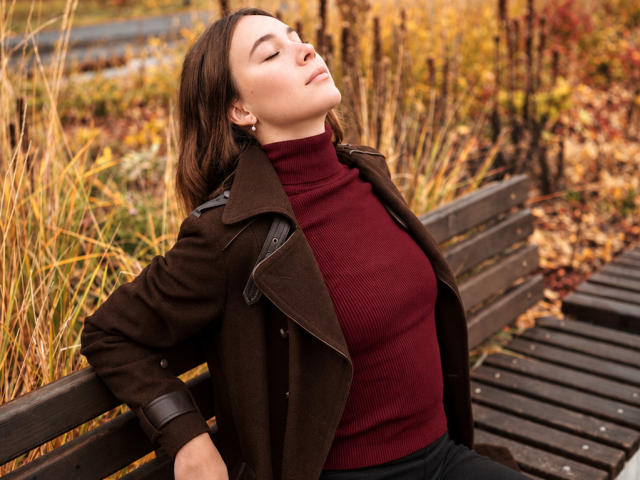 AmmaLoyXX with closed eyes, brown coat, red turtleneck, and black pants, leaning back on a wooden bench, autumn leaves, and grass in the background, looking relaxed and content.