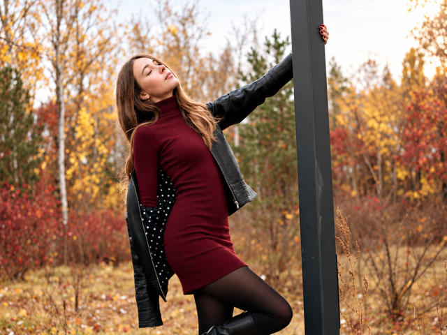 AmmaLoyXX, con los glúteos tensos, vestida con un vestido rojo de punto, leggings negros y una chaqueta de cuero, apoyada en un poste, con los ojos cerrados, cabello castaño largo, fondo de bosque otoñal..