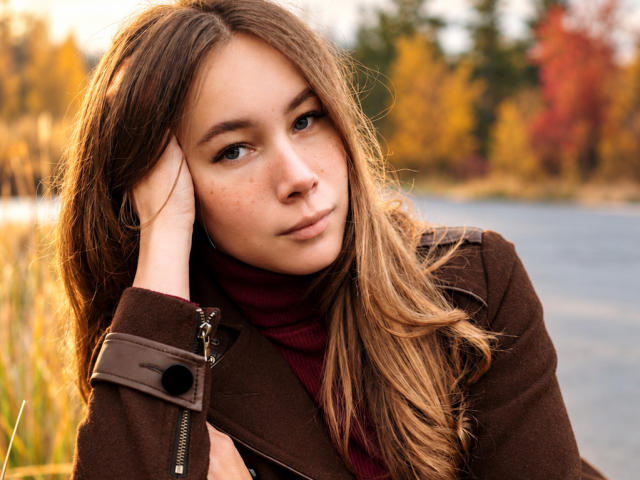 AmmaLoyXX, con el pelo largo y castaño, pecas y una expresión seria, vestida con un abrigo marrón y un cuello alto rojo, apoya la cabeza en la mano junto a un lago..