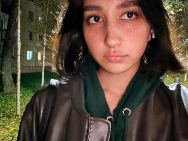 HeikeCarchi with medium brown skin, freckles, and dark brown wavy hair, wearing a green hoodie and brown leather jacket, has a lip piercing and a serious expression.