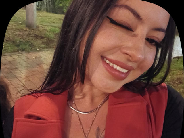 MiiaBennet has dark hair, is winking, and smiling with red lipstick. She's wearing a red shirt, black jacket, and two necklaces. Her tits are partially visible.