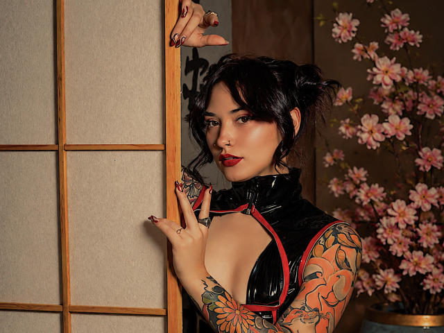 AkiraNishimura69 con una blusa de látex negro, mostrando el escote, tatuajes en los brazos, y sosteniendo el marco de una puerta, tiene lápiz labial rojo, cabello oscuro en mechones, y flores de cerezo rosadas en el fondo..