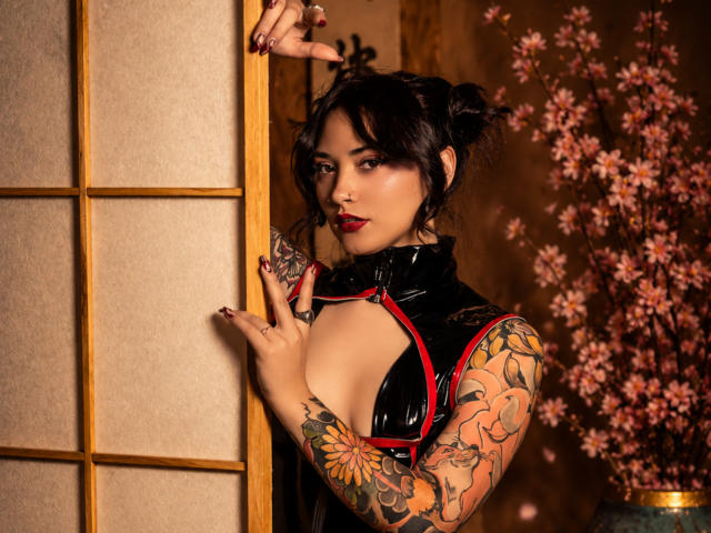 AkiraNishimura69, com tatuagens, vestindo uma blusa de látex preta, expondo os seios, batom vermelho, cabelo escuro em um coque bagunçado, segurando uma divisória de madeira, flores de cerejeira ao fundo..
