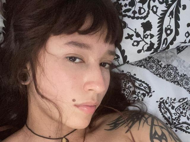 Julieth Courtouis mit dunklen, welligen Haaren, braunen Augen, einem kleinen Muttermal auf ihrem Kinn, schwarzen Ohrstöpseln, einem Tattoo an ihrer Schulter und einer schwarzen Halskette, liegt auf einem schwarz-weißen Kissen..